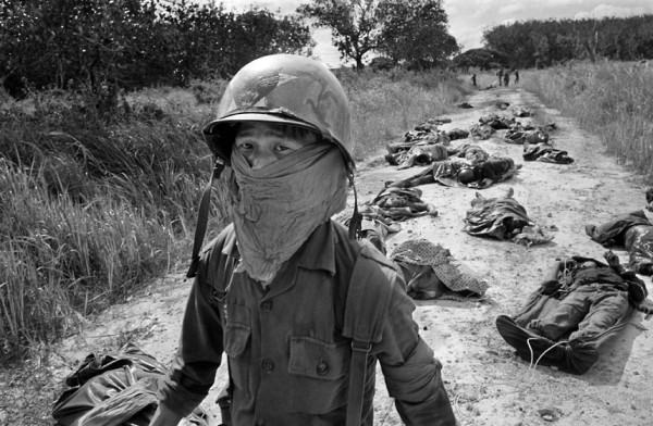 Bức ảnh được chụp vào tháng 11 năm 1965. Một người gom rác Việt Nam đeo mặt nạ để tránh mùi khi đi qua binh lính của Mỹ và binh sĩ Việt Nam bị chết trong cuộc giao tranh tại các đồn điền cao su Michelin, khoảng 45 dặm về phía đông bắc Sài Gòn.