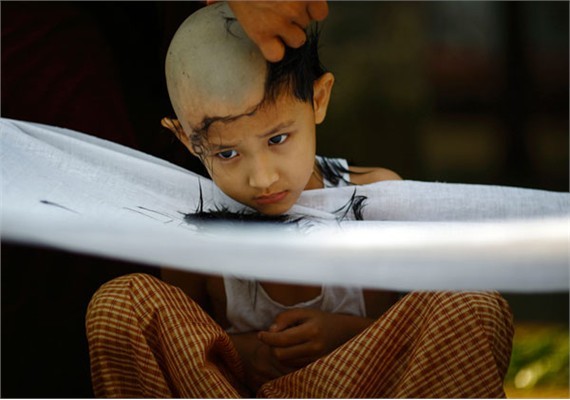 Cậu bé đang được một nhà sư cạo tóc. Theo truyền thống, tất cả các cậu bé ở độ tuổi từ 7 – 18 ở Myanmar đều được gia đình gửi vào chùa và sống cuộc sống tu hành trong 7 ngày.