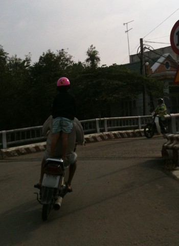 "Trẻ em làm xiếc cùng ba": Em bé một tay giữ mũ, còn một tay bám lấy ba trên xe máy. Chụp tại thị trấn Lái Thiêu, Thuận An, Bình Dương. Ảnh: Trịnh Văn Hải.