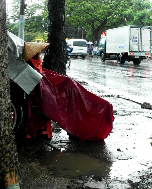 Trong cơn mưa tầm tã, người phụ nữ vá xe vỉa hè này trùm áo mưa, đưa đôi mắt buồn nhìn dòng đời xuôi ngược.