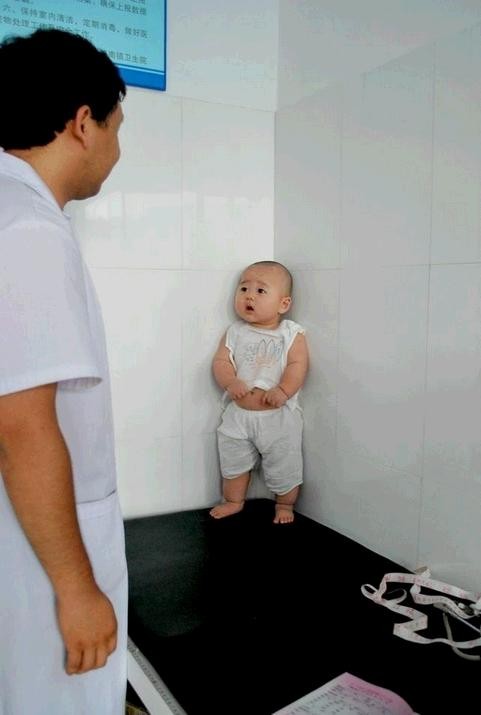 Cặp lông mày rướn cong, hai tay có vẻ phòng vệ, bé con đứng dán vào góc tường trên giường bệnh khi chuẩn bị được tiêm phòng.