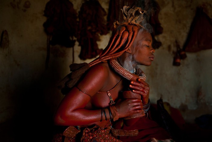 Những người phụ nữ Himba ở bắc Namibia thực hiện một nghi lễ hàng ngày bằng cách xông hỗn hợp đất son, dầu và tro lên người để bảo vệ cơ thể khỏi thời tiết khắc nghiệt của sa mạc. Họ không bao giờ tắm, nhưng sẽ đốt cây cỏ thơm trong một chiếc lọ vào mỗi sáng và xông lên người như là xức nước hoa. Ảnh: Dominique Brand.