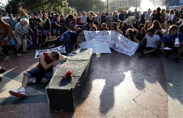 Một người biểu tình giả vờ khóc cạnh chiếc quan tài giả đại diện cho cái chết của nền giáo dục công trong cuộc biểu tình của phong trào Indignados tại Barcelona. Trên cỗ quan tài viết dòng chữ "các trường đại học công" ngày 14/5.