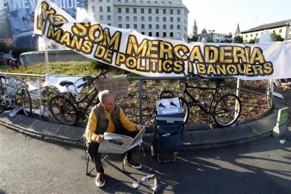 Một người đàn ông ngồi đọc báo dưới biểu ngữ "hàng hóa không nằm trong tay các chính trị gia và chủ ngân hàng" của phong trào biểu tình Indignados tại Barcelona ngày 14/5.